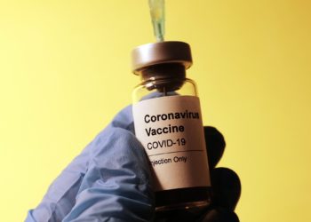 Image displays doctor/nurse extracting COVID-19 vaccine from vial. The hands are wearing blue sanitary gloves.