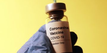 Image displays doctor/nurse extracting COVID-19 vaccine from vial. The hands are wearing blue sanitary gloves.