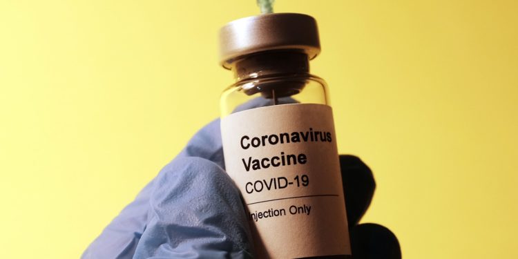 Image displays doctor/nurse extracting COVID-19 vaccine from vial. The hands are wearing blue sanitary gloves.