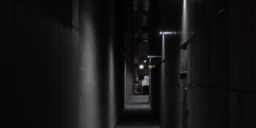 The image depicts a dark alley with several doorways on the right. Each doorway is lit by a dull lamp, which is mounted above it.