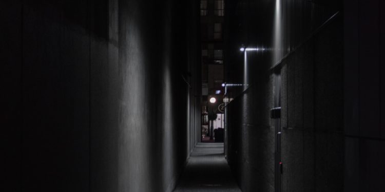 The image depicts a dark alley with several doorways on the right. Each doorway is lit by a dull lamp, which is mounted above it.