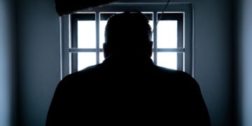Silhouette of a man facing a barred window.