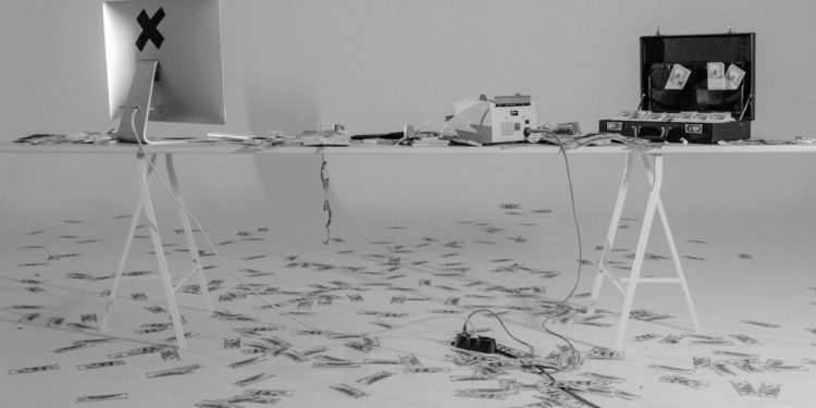 black and white image of a macbook computer screen - from the back - and an open suitcase. There are dollar bills all over the floor