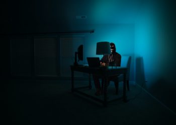 Dark room with desk in corner with person sat at it, lit by blue screen light.