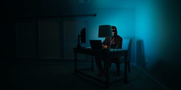 Dark room with desk in corner with person sat at it, lit by blue screen light.