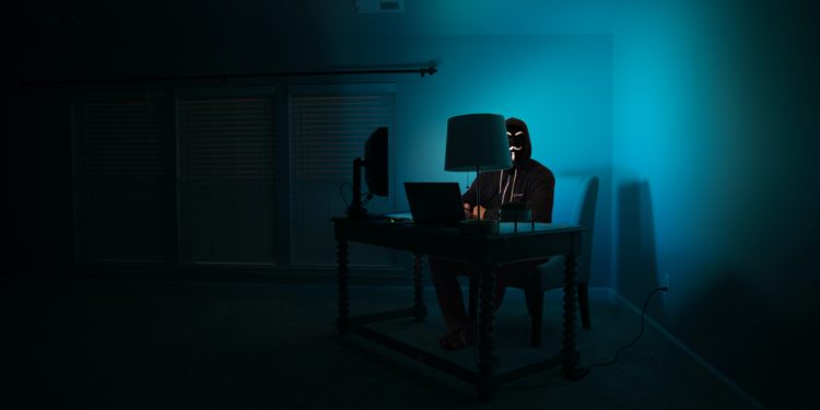 Dark room with desk in corner with person sat at it, lit by blue screen light.