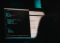 Two computer screens filled with code. Shadowed figure.